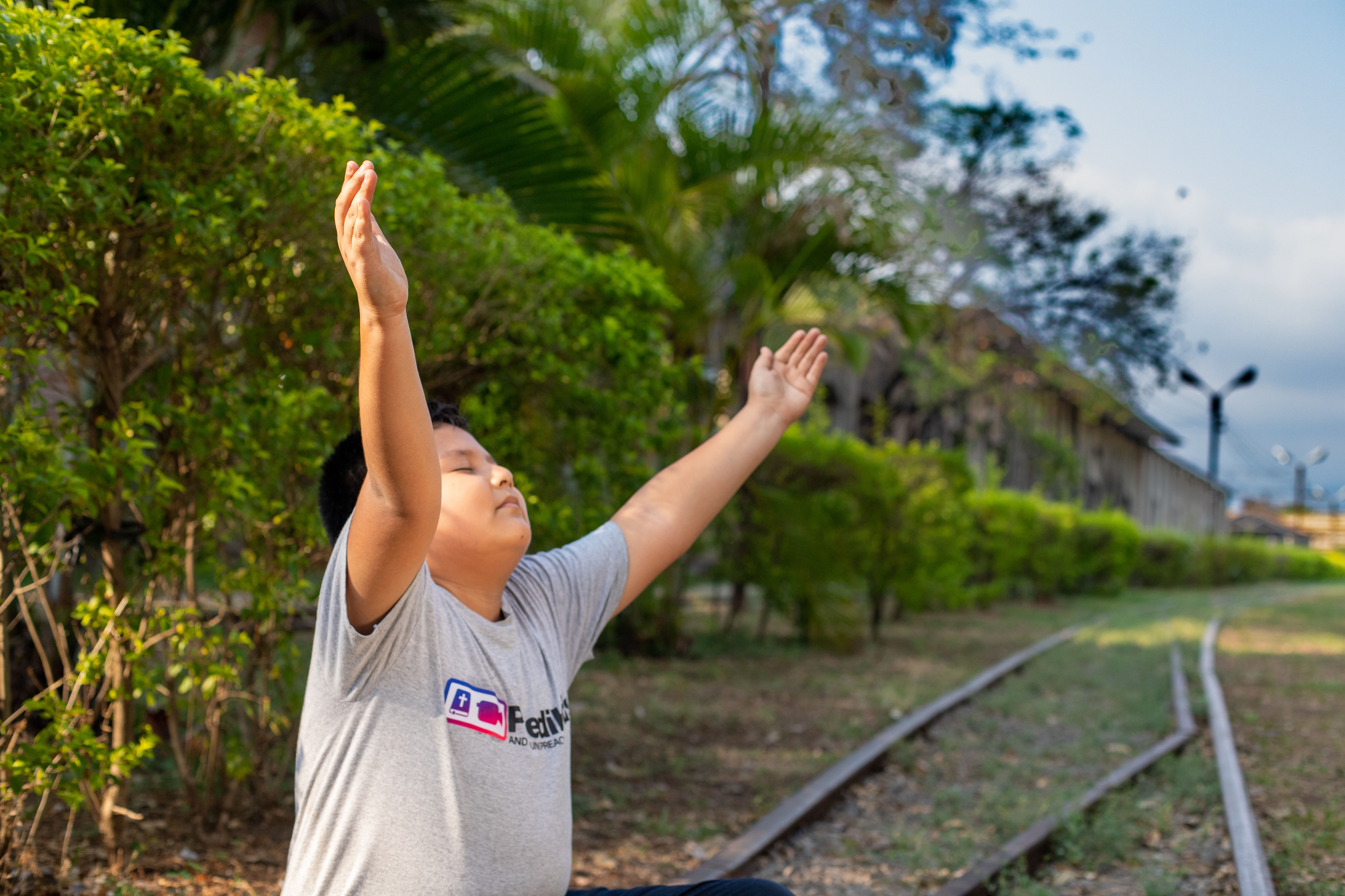 Young boy wearing a gray t-shirt sits on railroad tracks and lifts his arms in the air with his eyes closed.