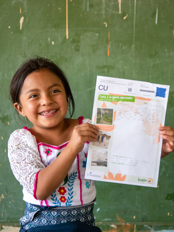 Lizbeth is wearing a white shirt with a floral pattern on it. She is standing in front of a green board in her classroom and she is holding up one of her sponsor's letters.