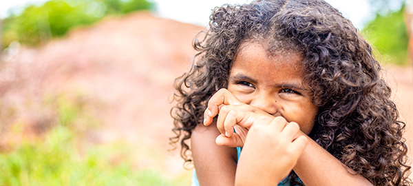 A young girl with curly hair covers her mouth with her wrist as she laughs